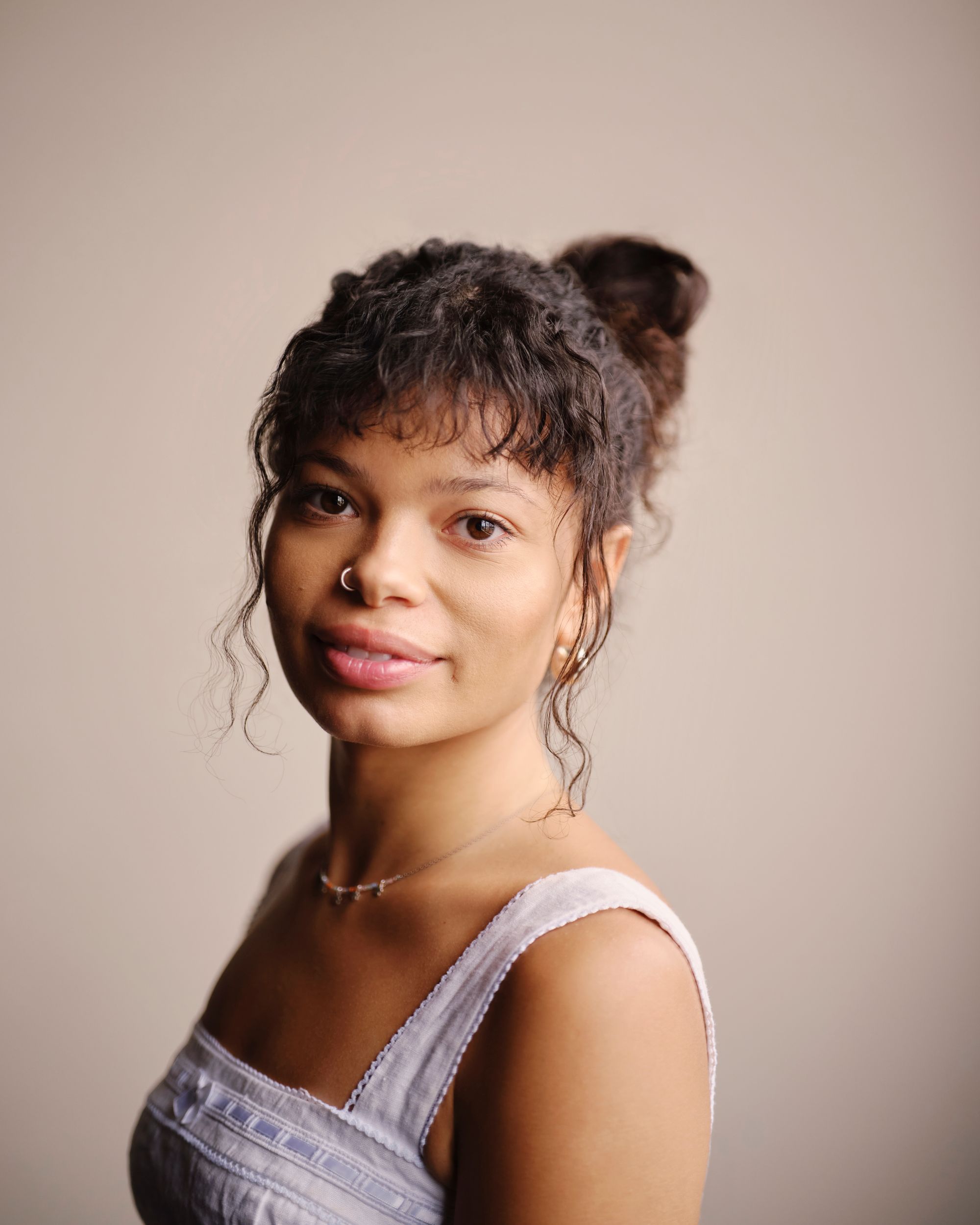 Beth, a twenty-something year-old Mixed-Race woman, is standing in front of a white wall. She is wearing a white vest top, nose ring and hair in a bun with a fringe.