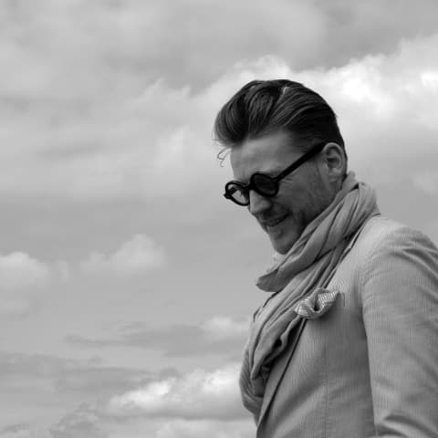 David is shown in a black-and-white photograph. He has short fair hair and is wearing a jacket, a large scarf and sunglasses. He is standing up, and the background is filled with clouds.\n