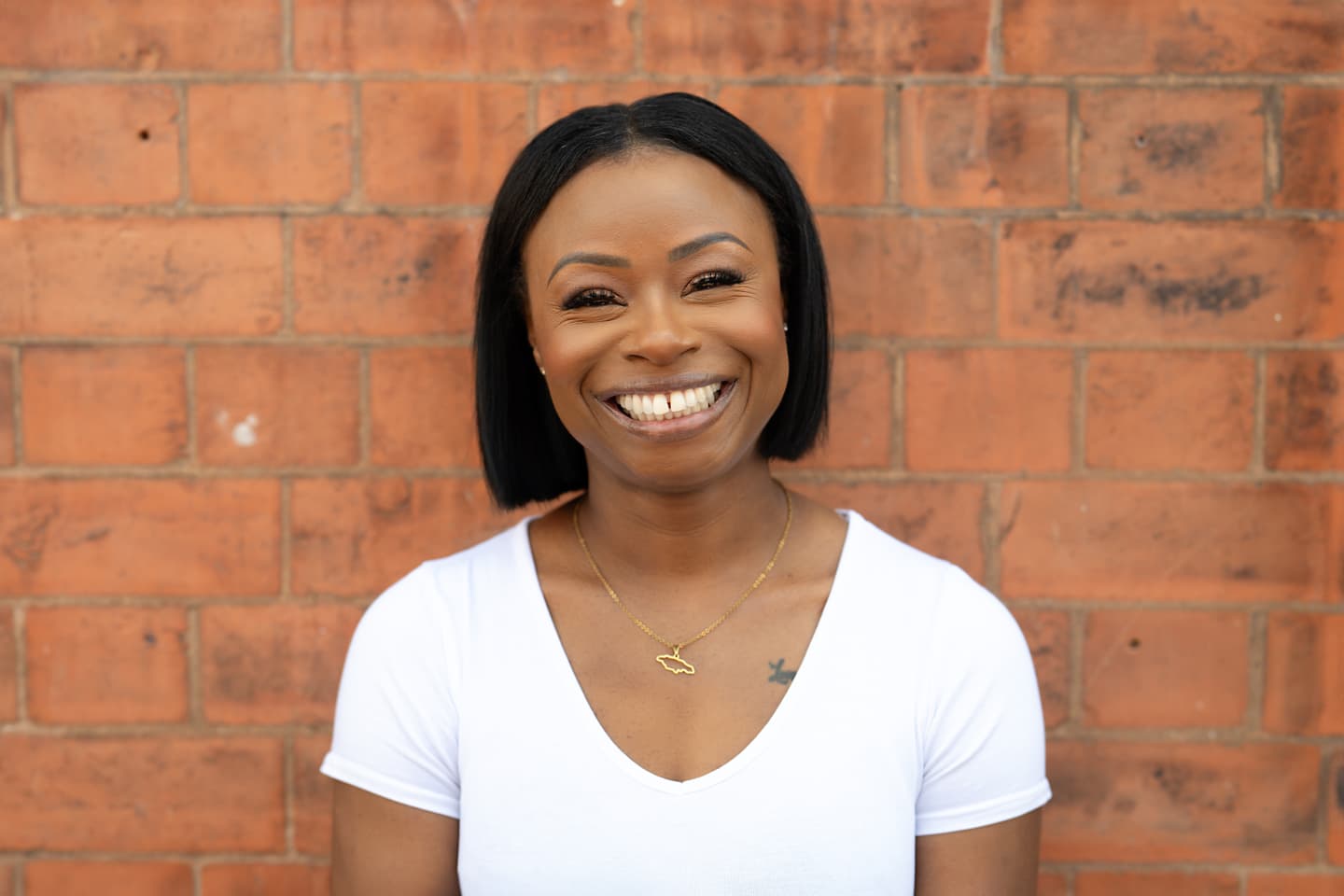 Liz is a Black woman with a big smile that shows her teeth. She has straightened black hair cut into a bob and is wearing a short-sleeved, v-neck white top with a delicate gold chain around her neck that has a small pendant on it in the shape of the island of Jamaica. She is standing outside, against an exposed brick wall.\n