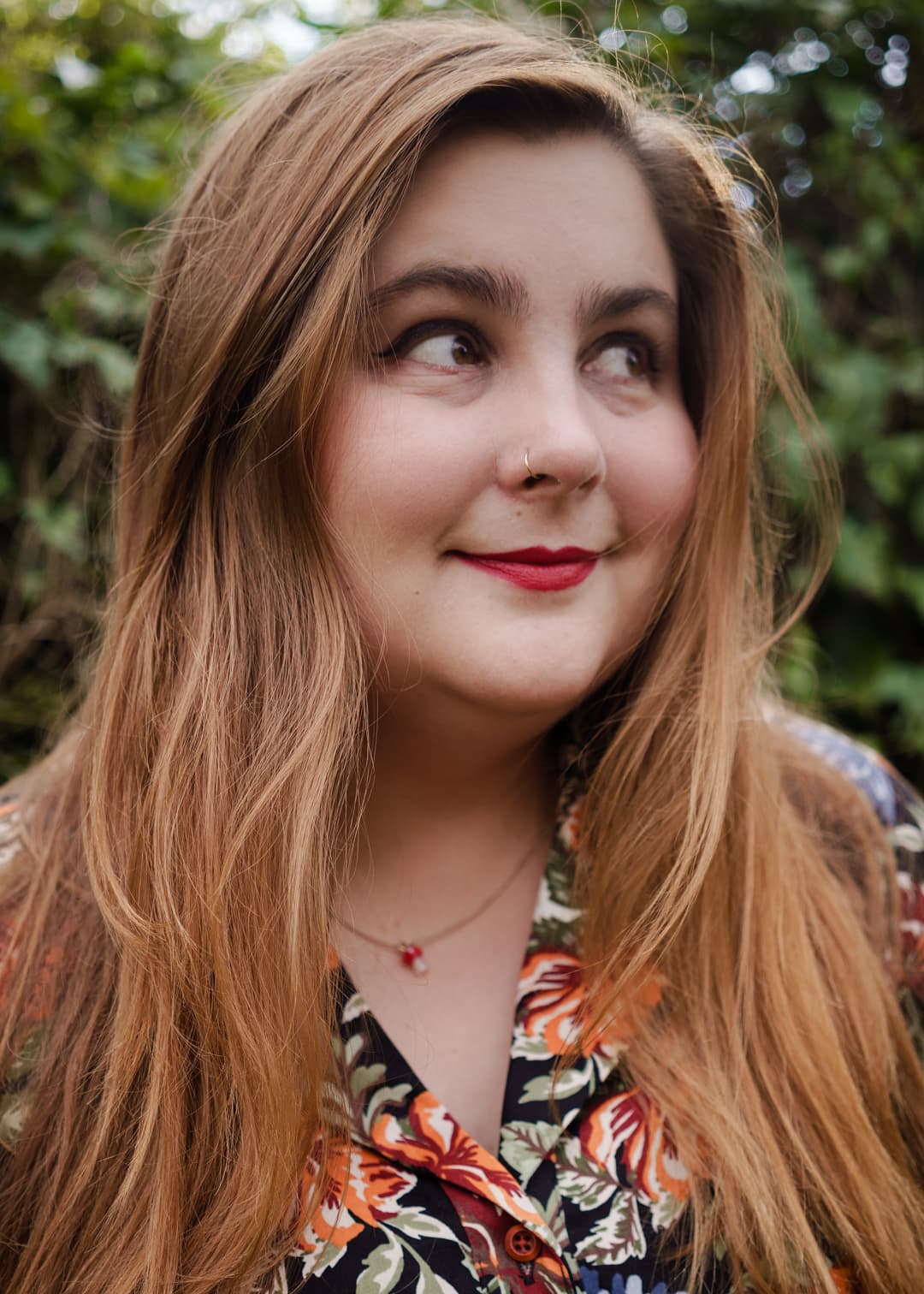 Lizzie is a white person with long light brown hair. They look away from the camera, with a cheeky smile. They wear red lipstick, a gold nose ring, a gold necklace with a mushroom pendant, and a rabbit print shirt dress.