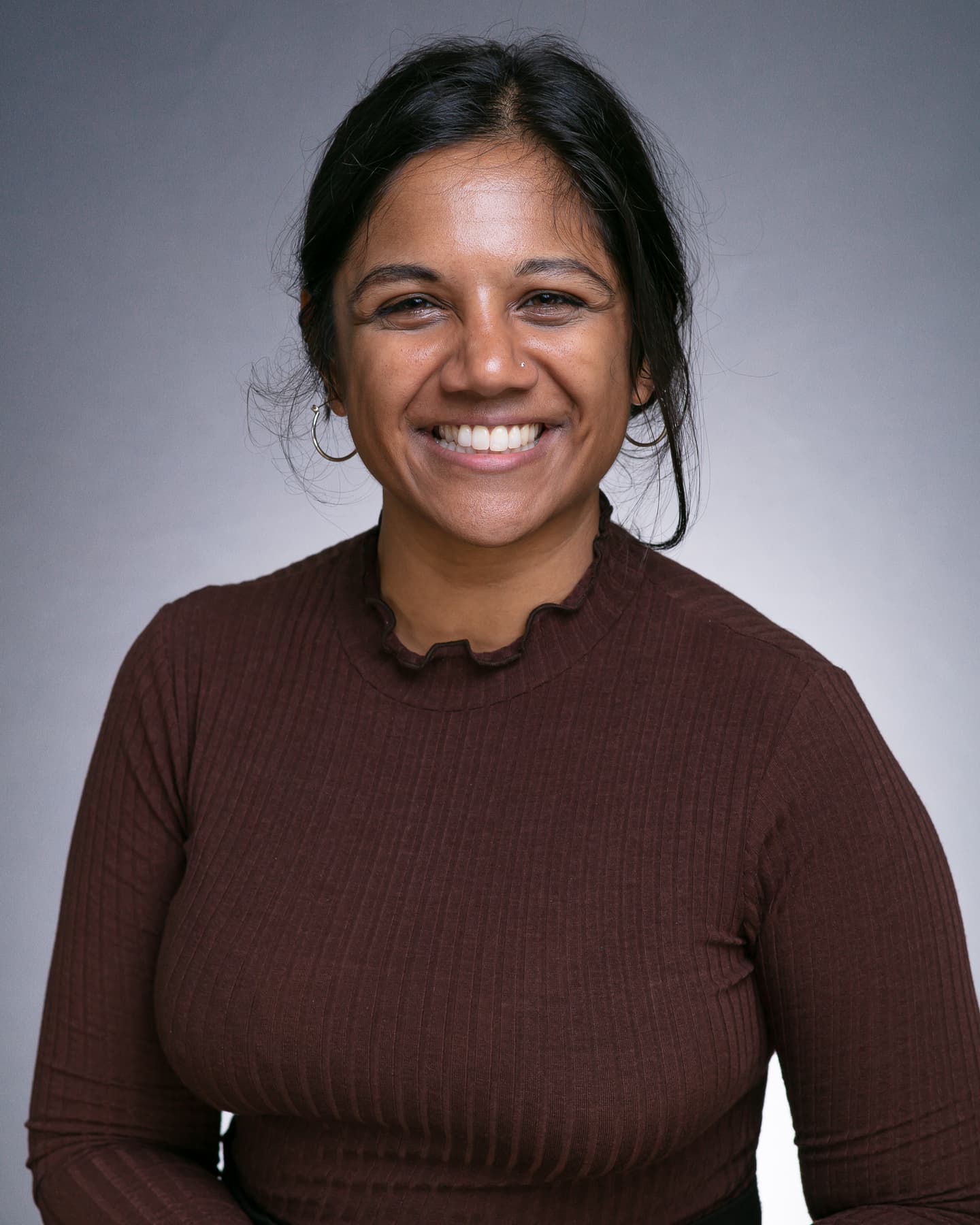 Rachna, a South Asian woman is sitting in front of a grey background. She is wearing a brown top, small silver hooped earrings and a small diamond in her nose. A small strand of her Black hair, which is tied back, falls out whilst she is smiling straight at the camera.