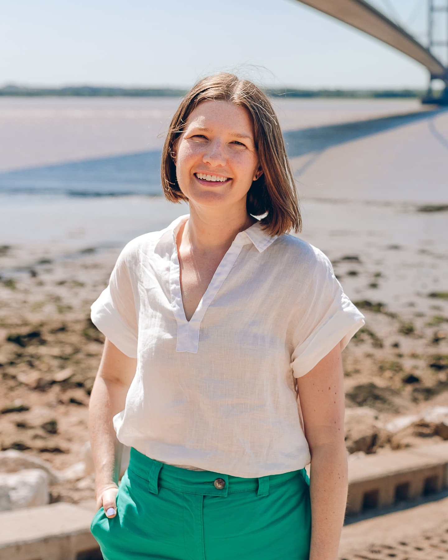 Sophie, a white woman with short, brown hair, is standing in front of a large suspension bridge. She is wearing a white shirt and green trousers and has her hand in her pocket.