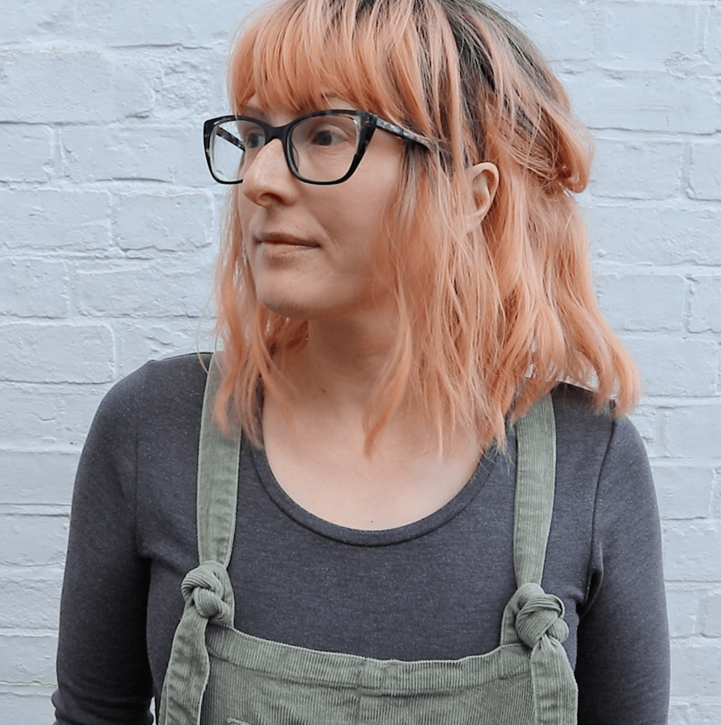 Jen, a thirty-something white woman, is wearing green dungarees, a grey top, glasses and a peach wig. She is standing in front of a white brick wall.