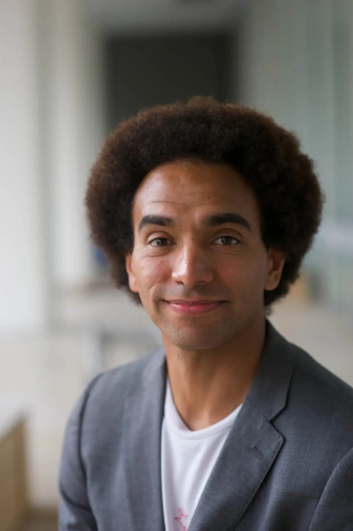 A headshot of former children's laureate, Joseph Coelho, a black bi-racial male with black curly hair styled as an afro. He is wearing a grey suit jacket