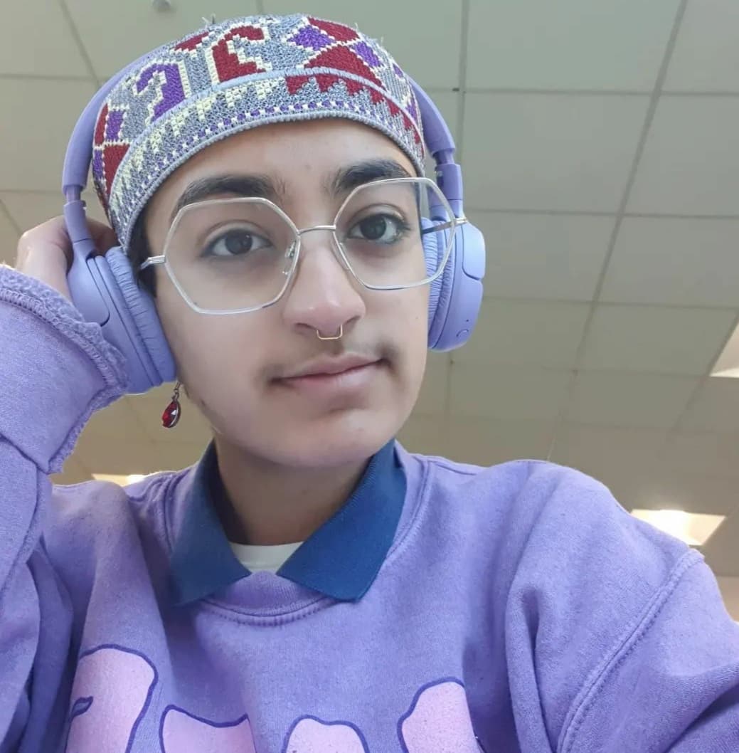 Moses is a brown person in their early twenties, slightly smiling under a white tiled ceiling. They are wearing a grey topi hat with red, purple, and cream patterns woven into it, under a pair of purple headphones. Their left hand rests on the left side of their headphones. Adorning their face is a pair of silver hexagonal framed glasses, golden square septum jewellery, and a red gem-drop earring on their left ear. They are wearing a purple jumper showing parts of a pink graphic, and a dark blue collar and white shirt poke out at the neckline.