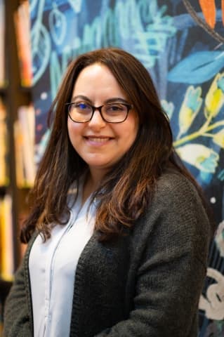 Farrah, a brunette with glasses, wearing a grey shirt and cardigan, standing in front of a bookshelf.