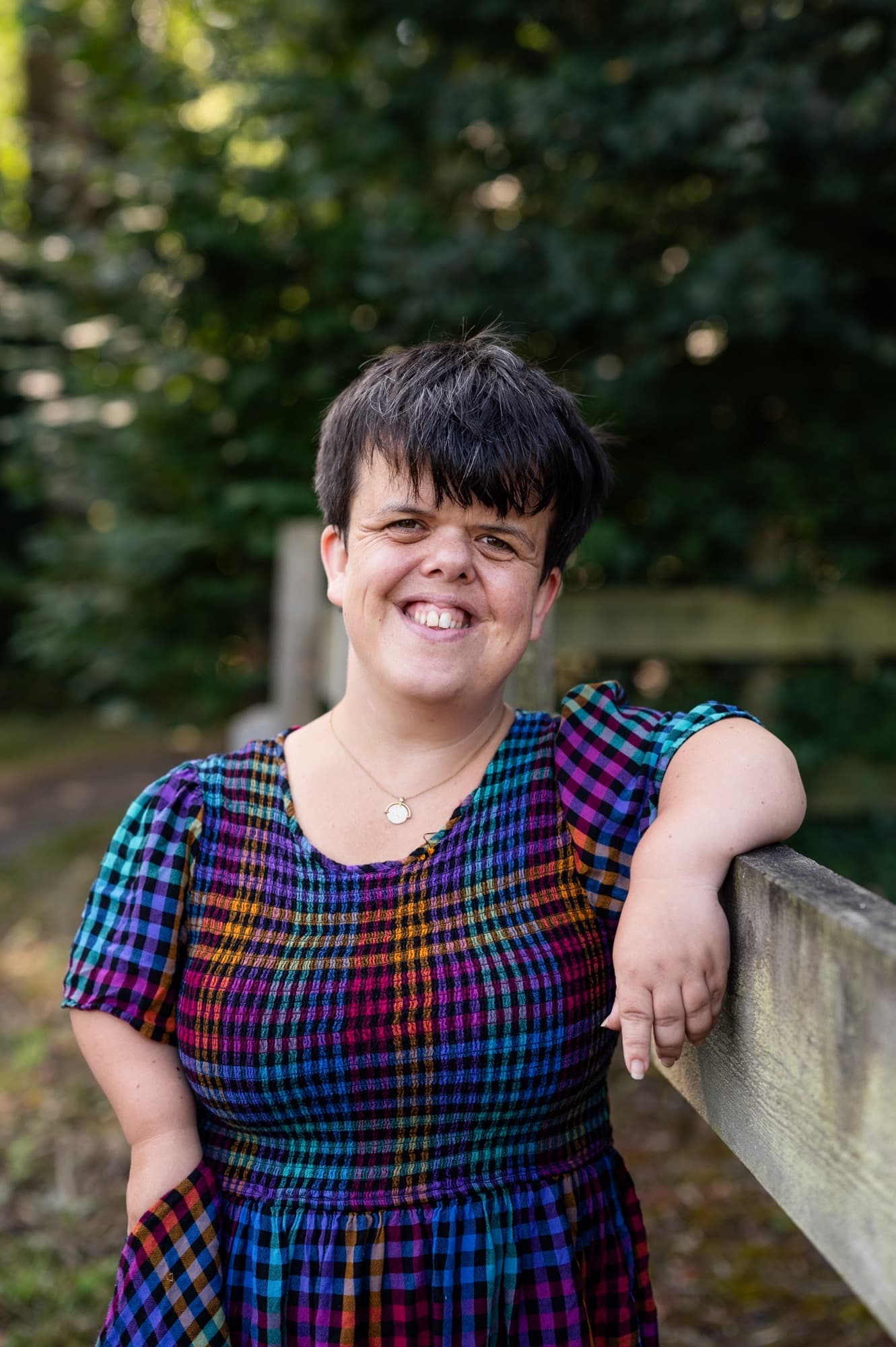 Profile shot of Cathy Reay, a white woman with dwarfism and short, dark-brown hair. She's wearing a multicoloured checked dress and leaning against a wooden fence. There are trees behind her and she’s smiling widely.